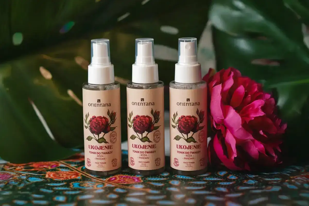 Trzy buteleczki toniku do twarzy Orientana z różą i pandanem, otoczone liśćmi i kwiatem piwonii.