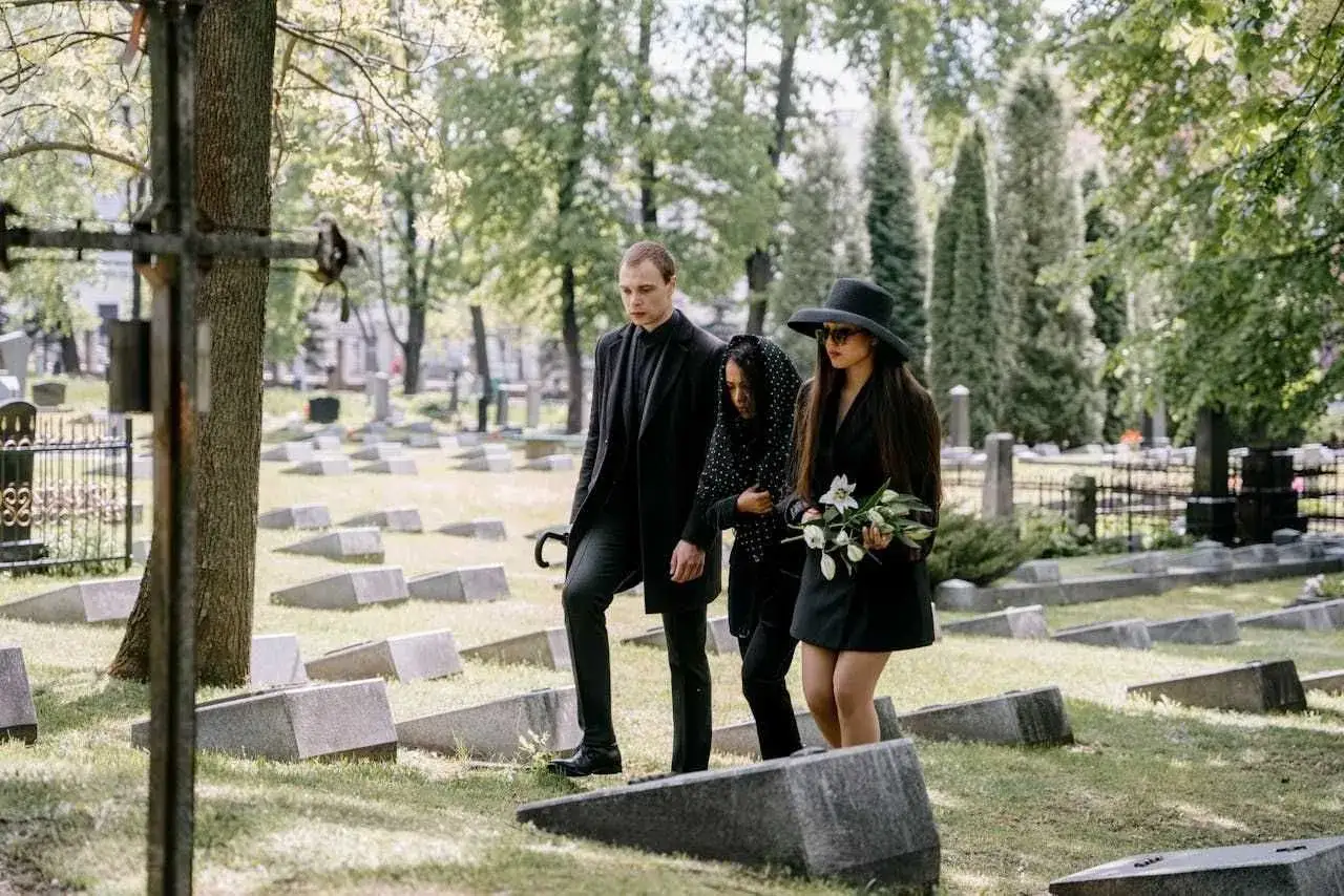 Trzy osoby w czarnych ubraniach idące przez cmentarz, jedna z bukietem kwiatów.