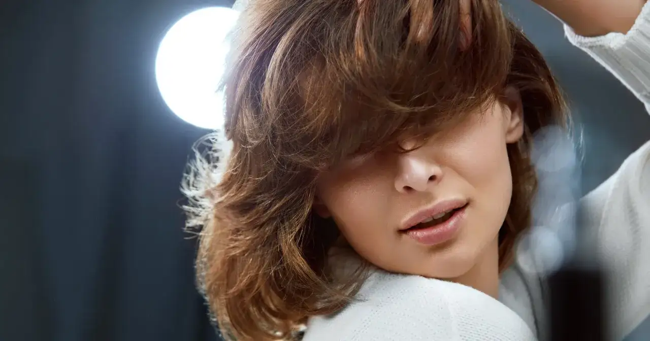 Kobieta z burzą brązowych włosów, zastanawiająca się, jak utrwalić fryzurę na wesele. Delikatne światło podkreśla jej urodę.