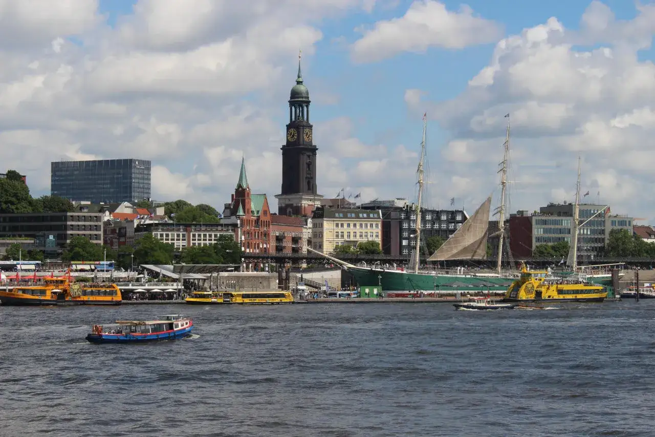 Widok na port w Hamburgu, gdzie statki i łodzie pływają po wodzie, a w tle widać miasto z wieżą kościelną. Czy Hamburg leży nad morzem? Tak, to miasto portowe!