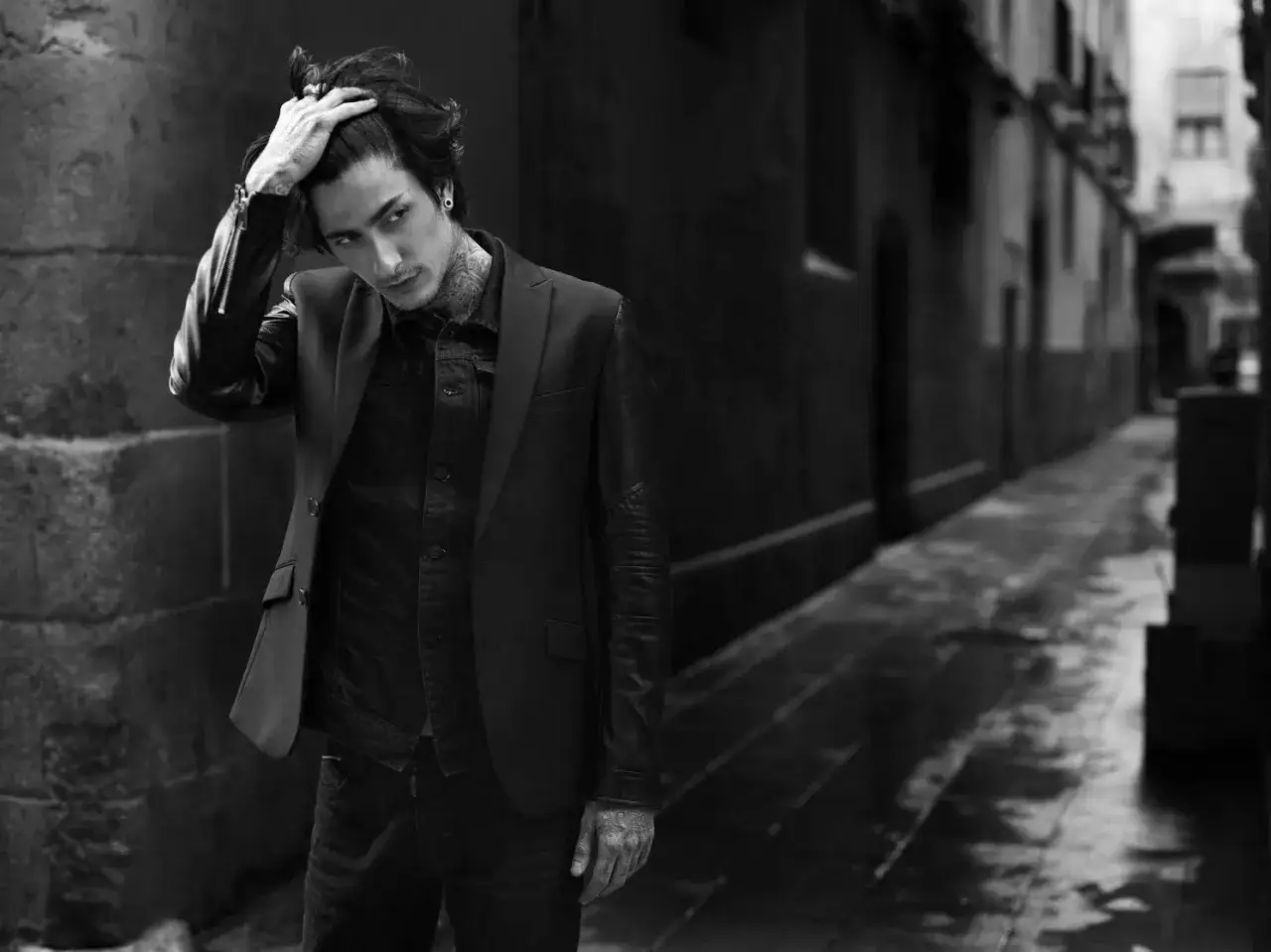 Młody mężczyzna w stylowej marynarce Antony Morato, z tatuażami, stoi w ciemnej uliczce.