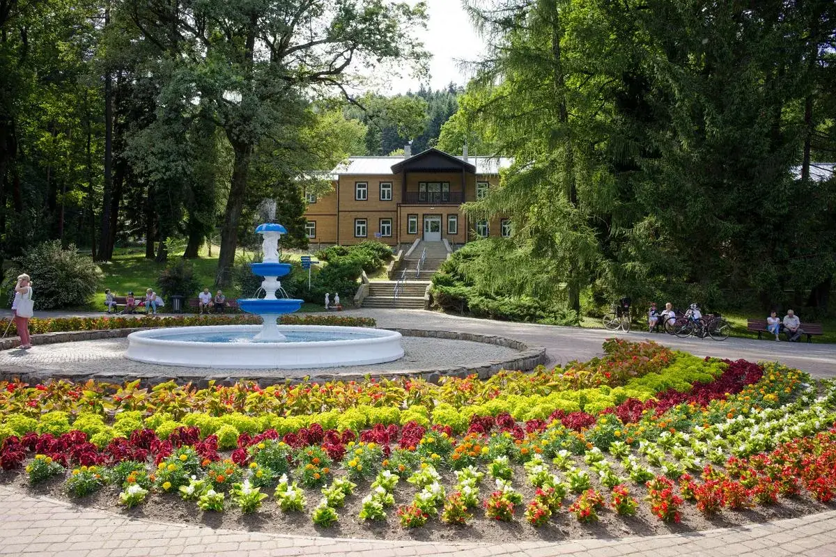 Uzdrowisko z fontanną i kolorowymi rabatami kwiatowymi przed budynkiem sanatorium.