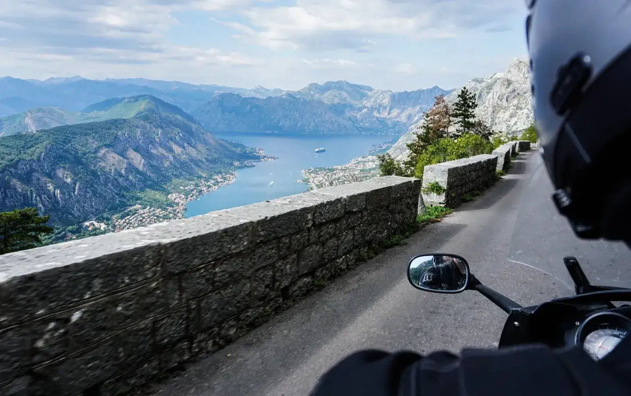 Widok z motocykla na Zatokę Kotorską w Czarnogórze. Kręta droga, kamienny mur i zapierająca dech w piersiach panorama gór i morza.