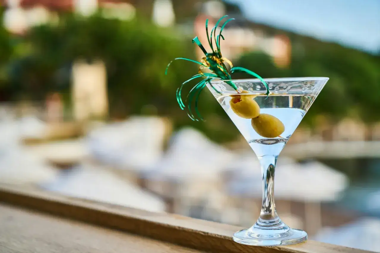 Idealny martini aperitivo z dwiema oliwkami, ozdobiony zielonymi wstążkami, na tle letniego krajobrazu.