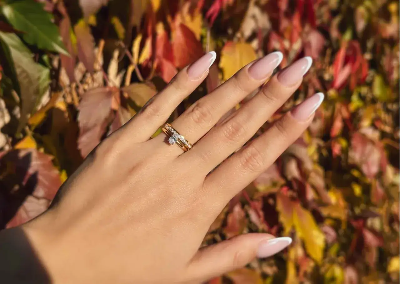 Dłoń z paznokciami w stylu francuskiego manicure i dwoma pierścionkami z różowego złota, ozdobionymi błyszczącymi kamieniami, na tle jesiennych liści.