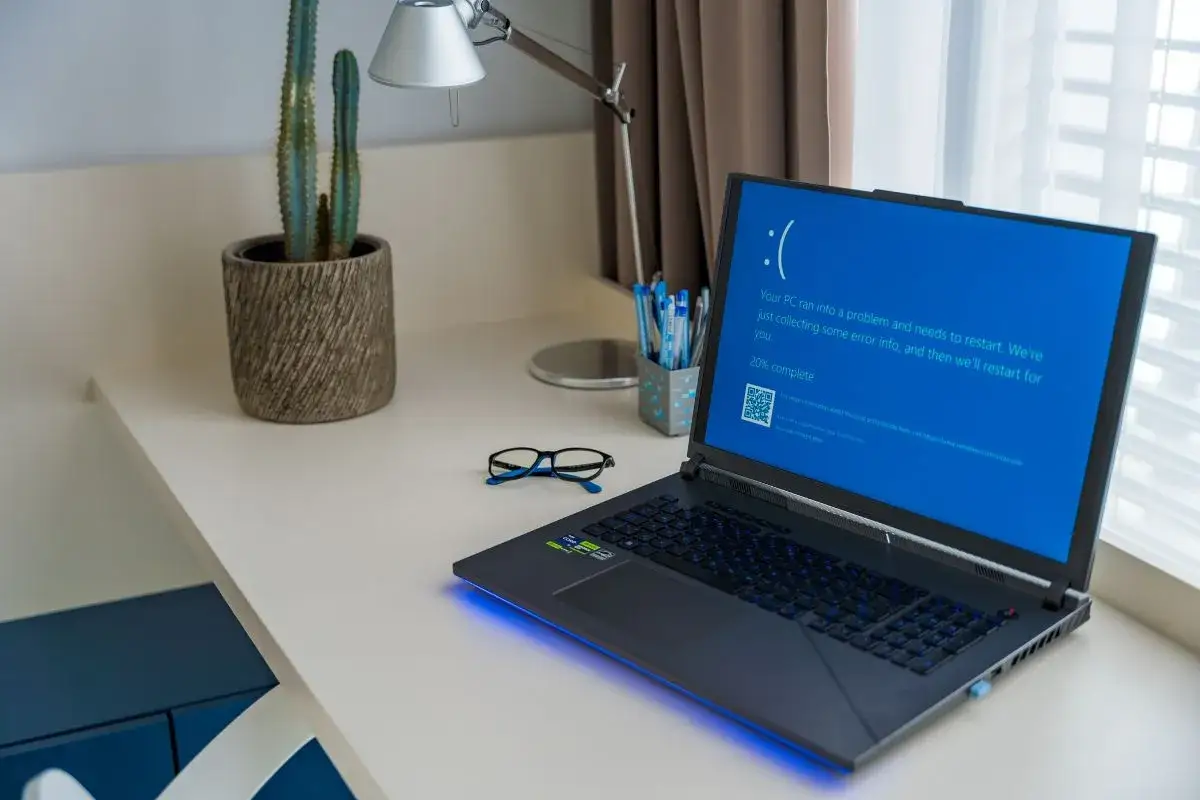 Laptop z niebieskim ekranem śmierci, wymagający twardy reset laptopa. Na biurku okulary i kaktus.