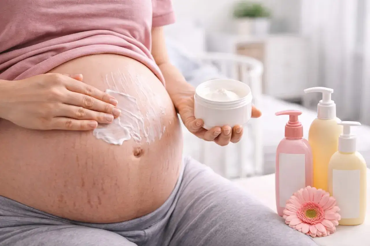 Ciężarna kobieta smaruje brzuch kremem. W pobliżu stoją butelki z kosmetykami i kwiat. Bezpieczne dla skóry w ciąży, np. bez kwasu glikolowego.