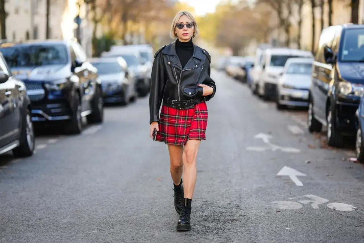 Kobieta w skórzanej ramonesce, czarnym golfie i czerwonej spódnicy w kratę prezentuje styl ubioru grunge. Na nogach ma ciężkie buty.