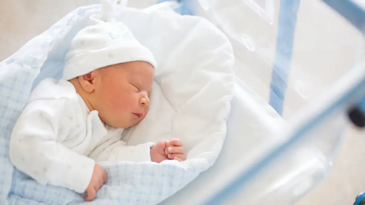 Noworodek śpi w łóżeczku szpitalnym, otulony białym kocykiem w niebieską kratkę.