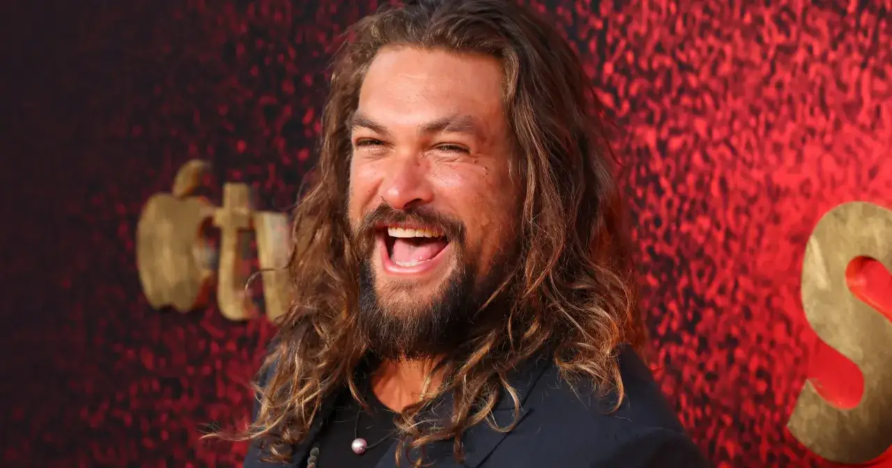 Jason Momoa śmieje się radośnie na tle czerwonej ściany z logo Apple TV+. Gwiazdor znany z wielu filmów, seriali i programów.