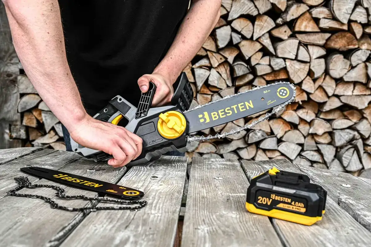 Ręce trzymają piłę łańcuchową Besten, przygotowując się do cięcia drewna. Obok leży zapasowy łańcuch i bateria.