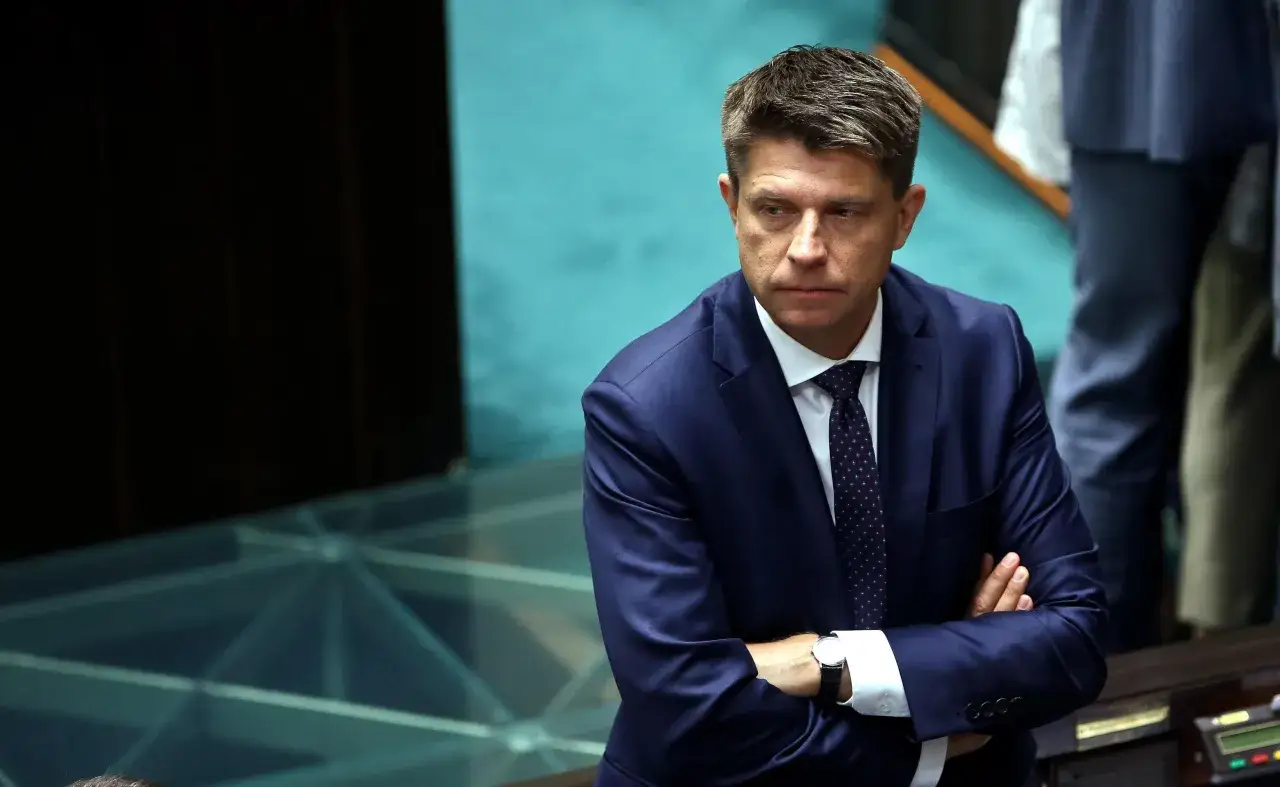 Ryszard Petru w garniturze z rękami skrzyżowanymi na piersi, w tle niebieskie siedzenia.