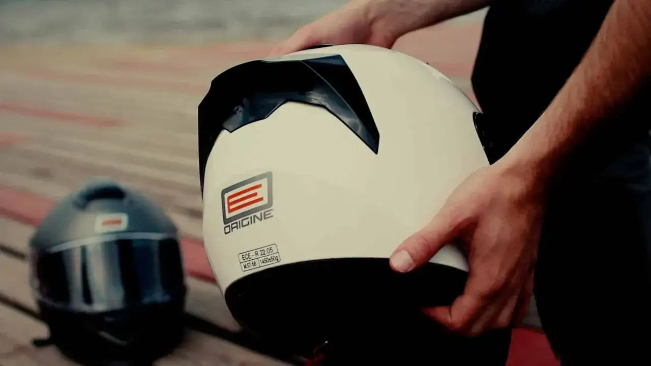 Ręce trzymają biały kask motocyklowy. Obok leży czarny kask. Dylemat: kask szczękowy czy integralny?
