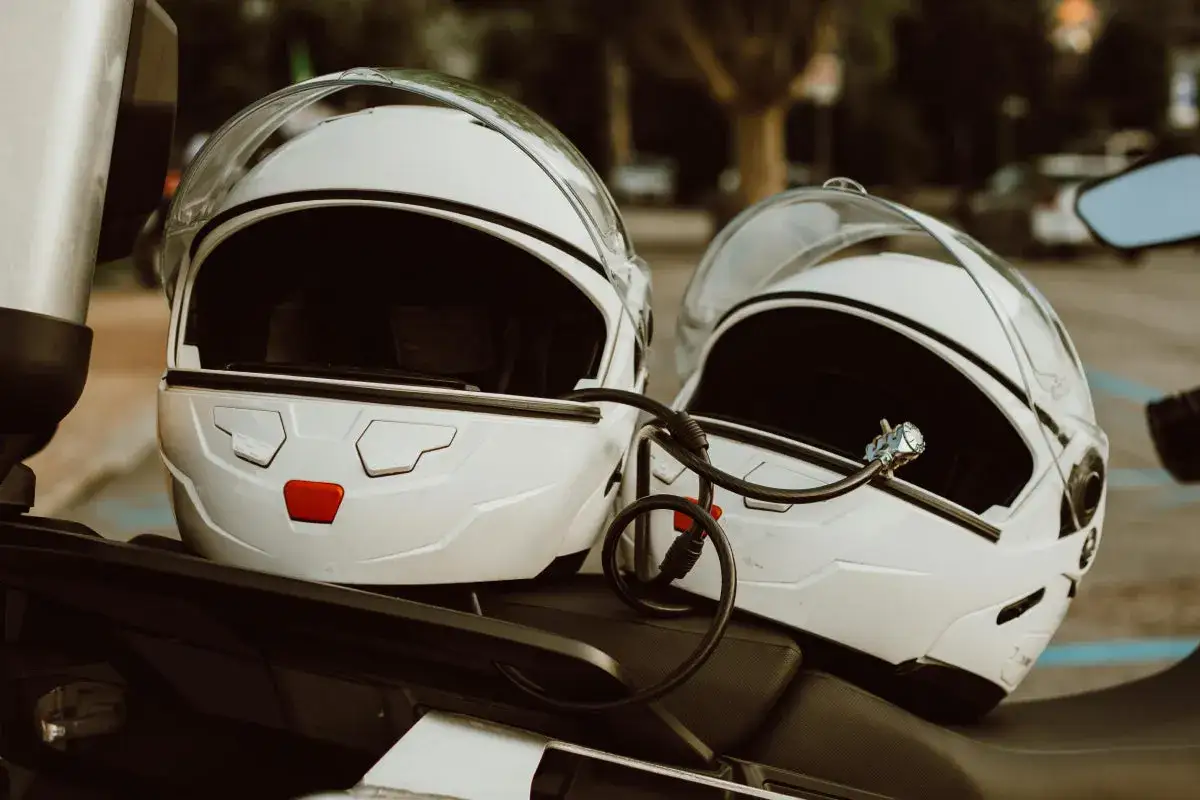 Dwa białe kaski motocyklowe połączone linką zabezpieczającą. Mocowanie kasku do motocykla zapewnia bezpieczeństwo podczas postoju.