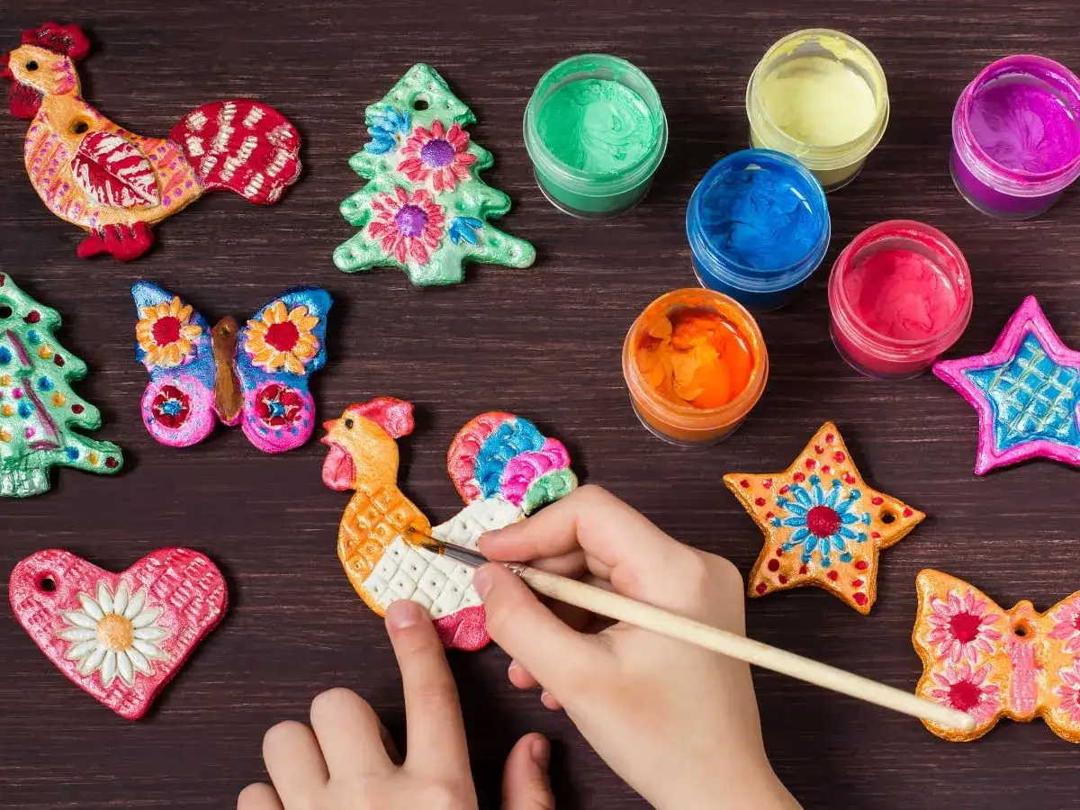 Dziecko maluje kolorowe ozdoby z masy solnej: koguty, choinki, gwiazdki i motyle.