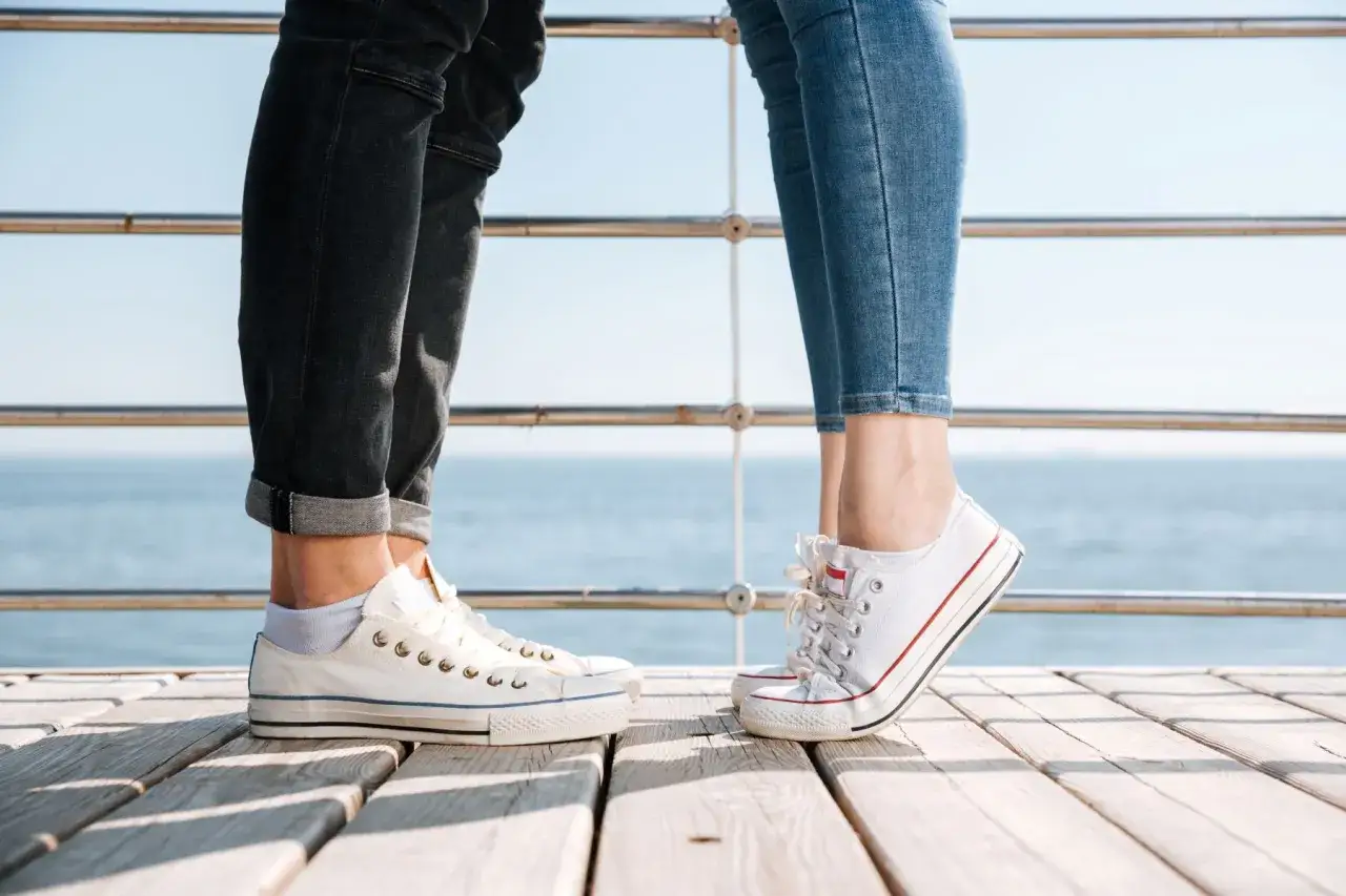 Para na drewnianym pomoście, w białych trampkach i jeansach. Idealne converse stylizacje damskie na letni dzień nad morzem.