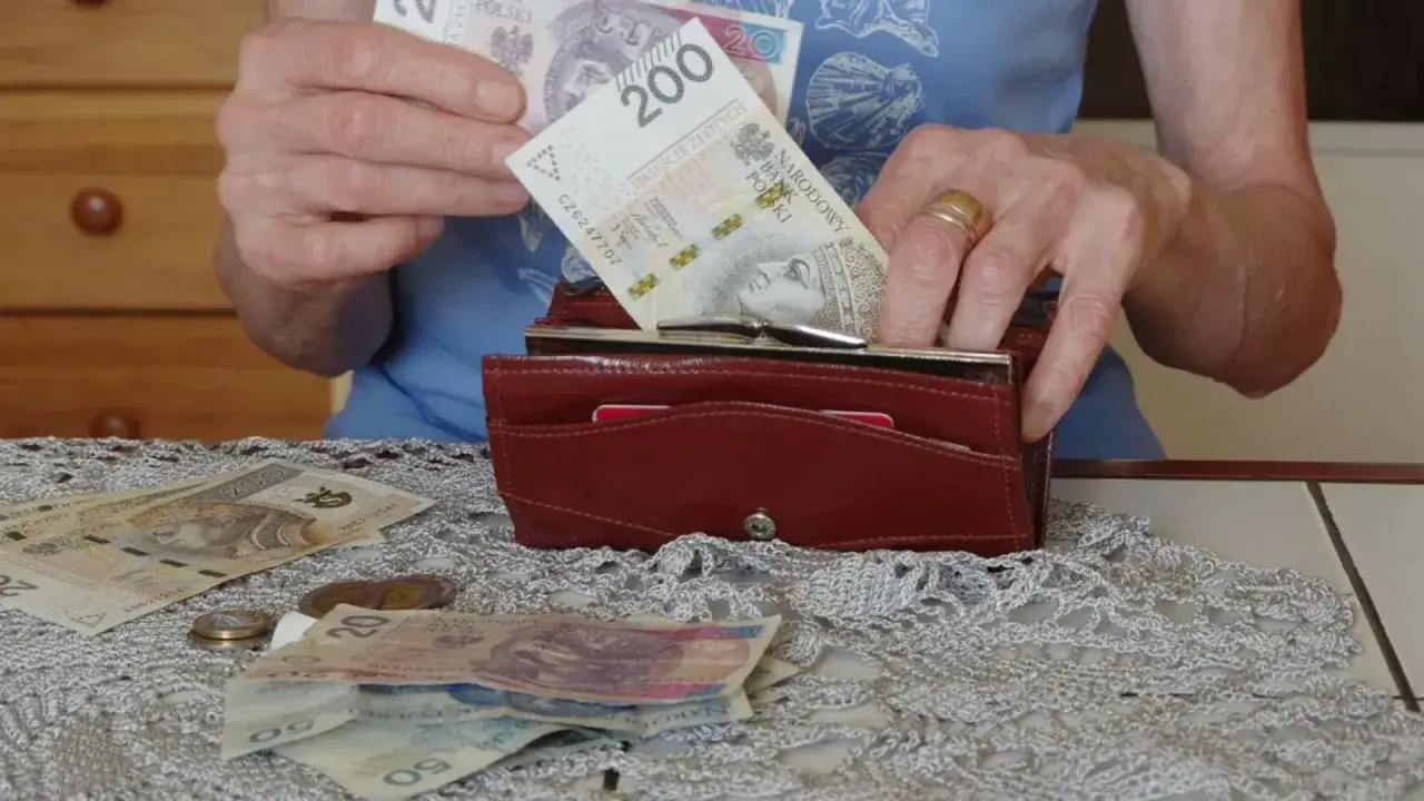 Dłonie wkładają banknot 200 zł do portfela. Na stole leżą inne banknoty i monety, co skłania do refleksji, ile wynosi średnia emerytura.