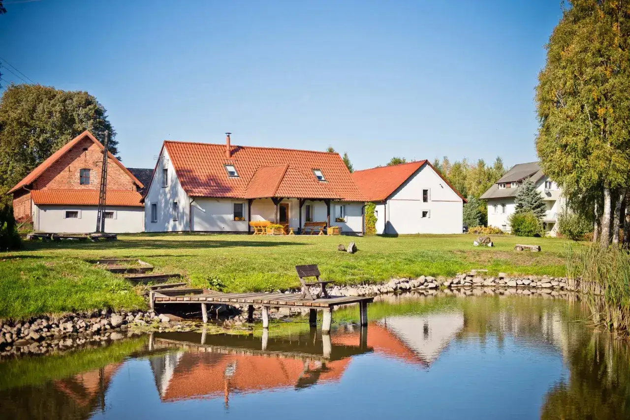 Dwa białe domy z czerwonymi dachami i staw z pomostem i ławką.