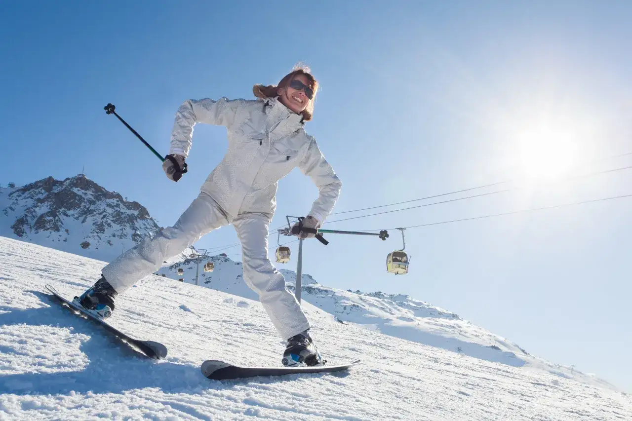 Uśmiechnięta narciarka w białym kombinezonie demonstruje, jak skręcać na nartach, z wyciągami i ośnieżonymi górami w tle.