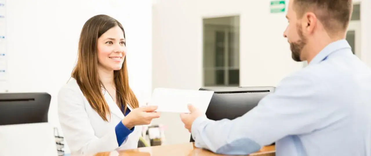 Uśmiechnięta recepcjonistka w białym fartuchu podaje pacjentowi dokumenty. Dowiedz się, jak sprawdzić wyniki badań.