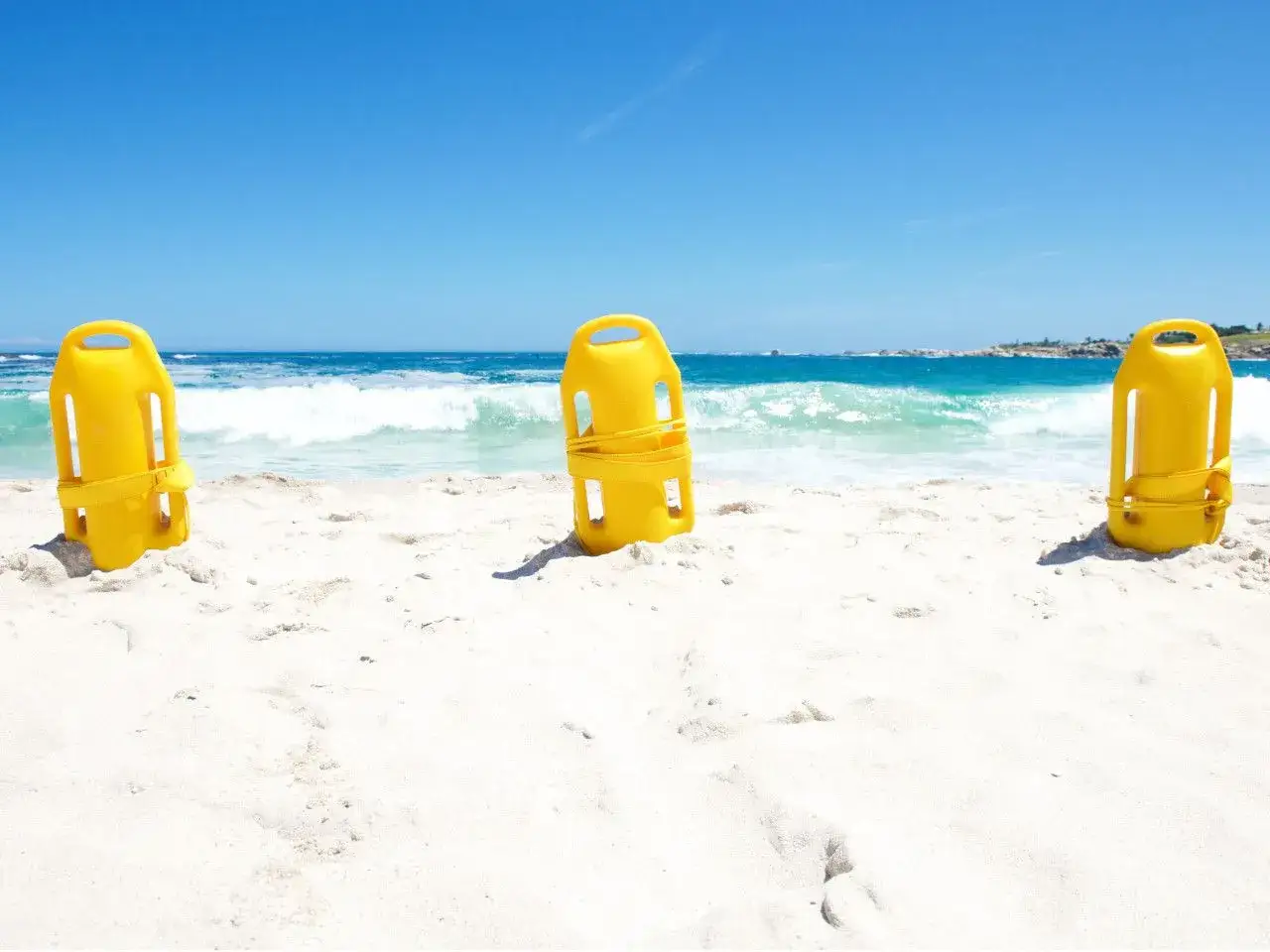 Trzy żółte bojki ratownicze stoją na piaszczystej plaży, z morzem i niebem w tle.