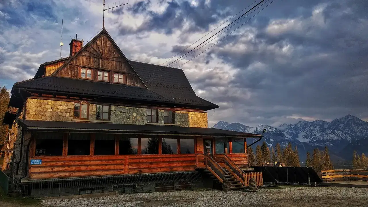Kamienno-drewniany dom na wierchu, z widokiem na ośnieżone szczyty gór.