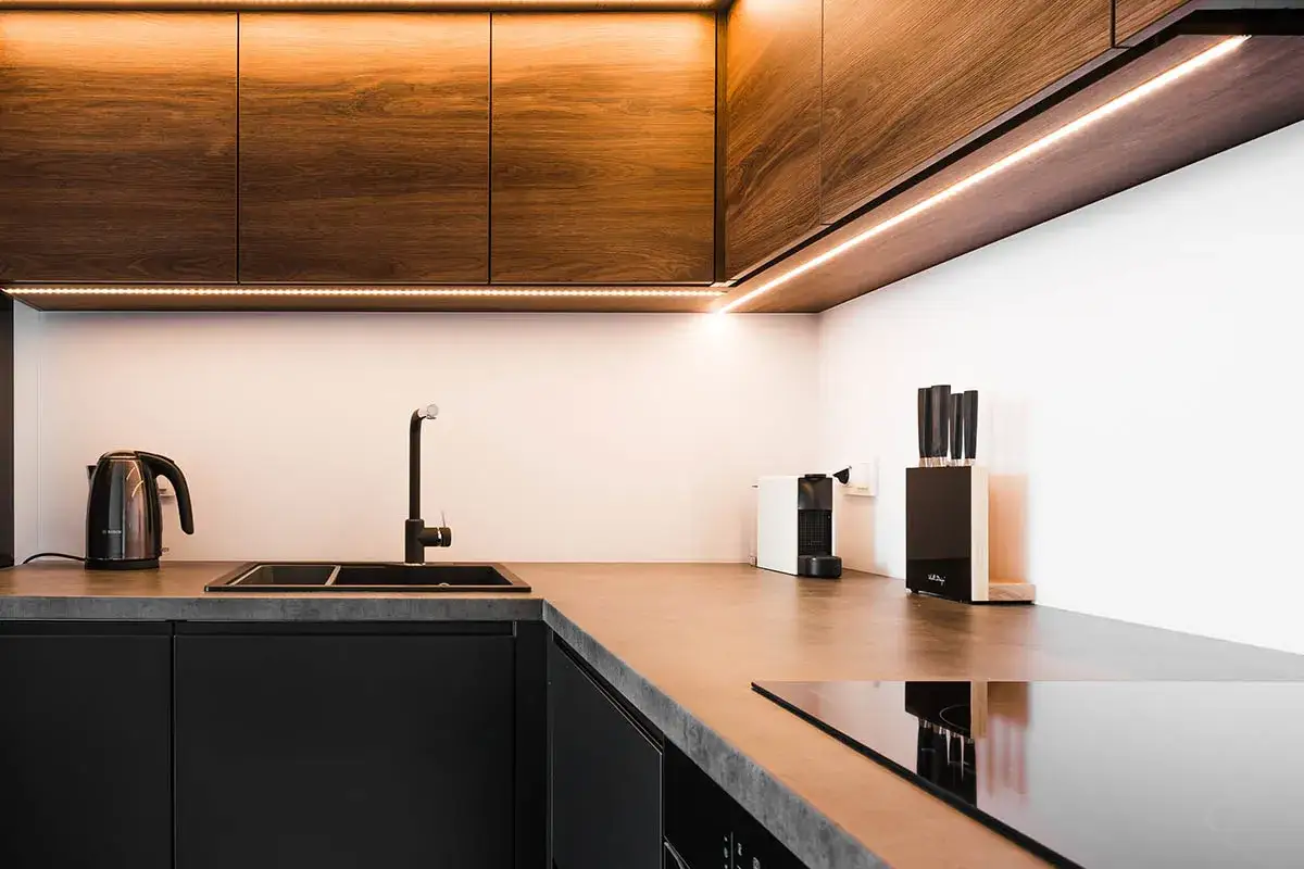 Nowoczesna kuchnia z drewnianymi frontami szafek i ciepłym oświetleniem LED pod szafkami kuchennymi. Czarny zlew, czajnik i ekspres do kawy.