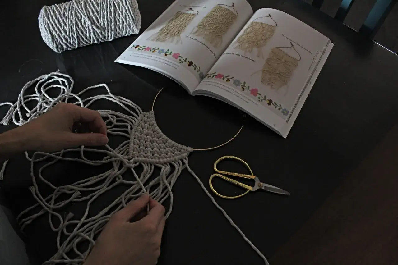 Ręce tworzą naszyjnik ze sznurka, obok leży książka z wzorami makramy i złote nożyczki.