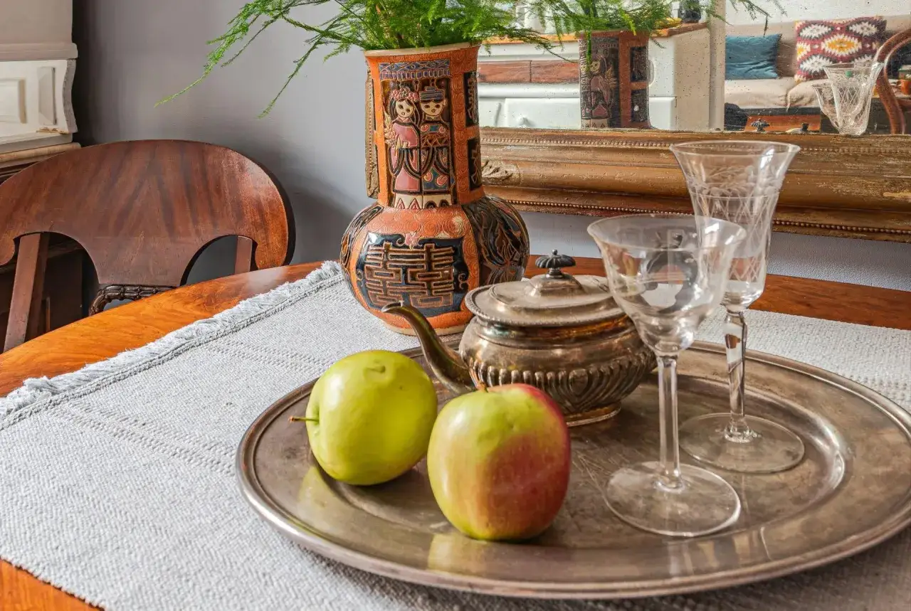 Srebrna taca dekoracyjna z jabłkami, czajniczkiem i kieliszkami. W tle wazon z roślinami i lustro.