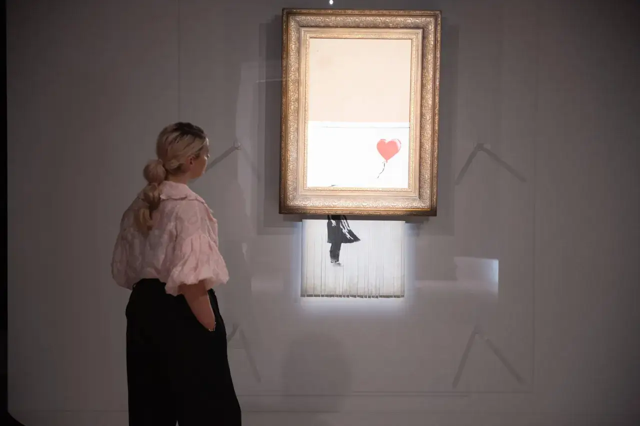 Kobieta ogląda słynne dzieło Banksy'ego, które uległo samozniszczeniu. Obraz z czerwonym balonem w kształcie serca jest częściowo zniszczony.