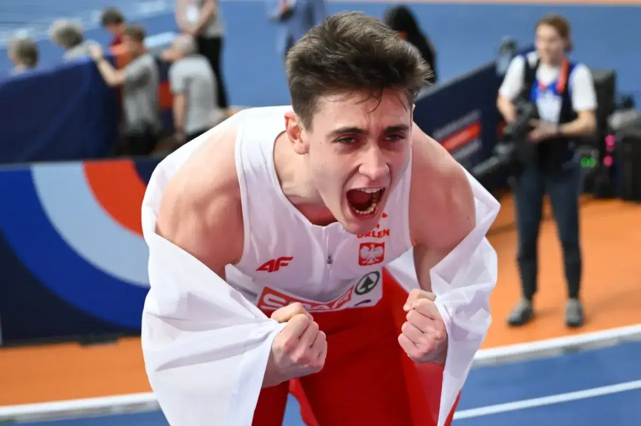 Młody lekkoatleta w biało-czerwonej koszulce okryty białą flagą, krzyczy z radości po zwycięstwie na halowych mistrzostwach europy lekkoatletyka.