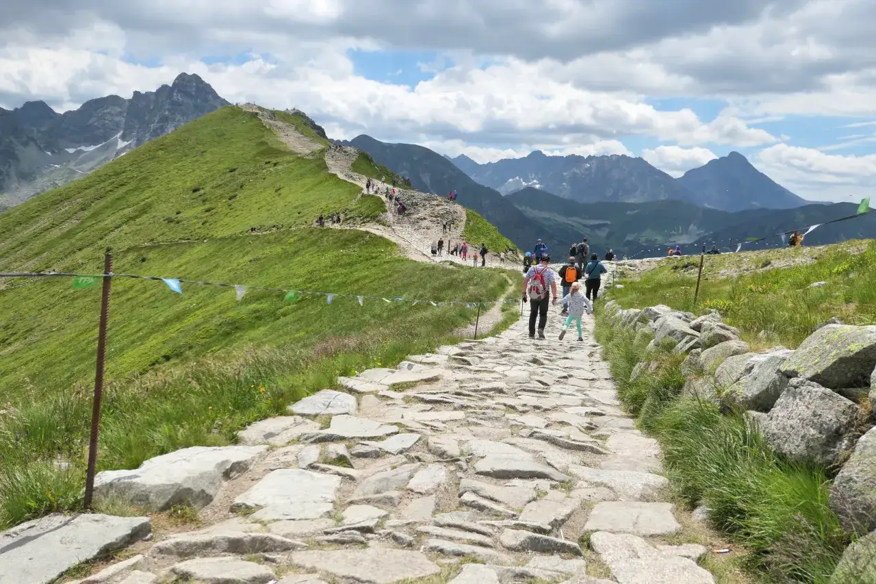 Kamienista ścieżka na zielonym zboczu górskim z turystami i widokiem na Tatry.