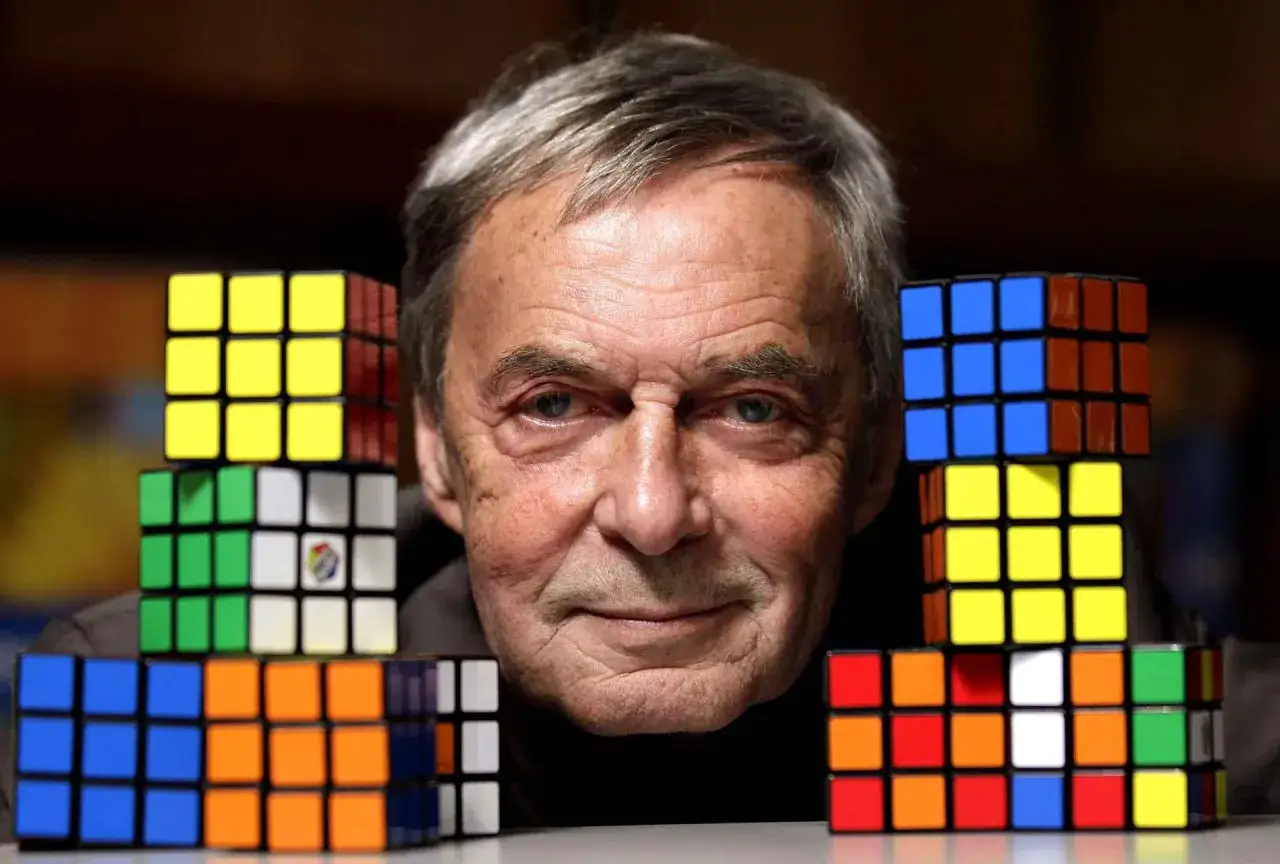 Erno Rubik, który wymyślił kostkę Rubika, z uśmiechem spogląda na stos kolorowych łamigłówek.