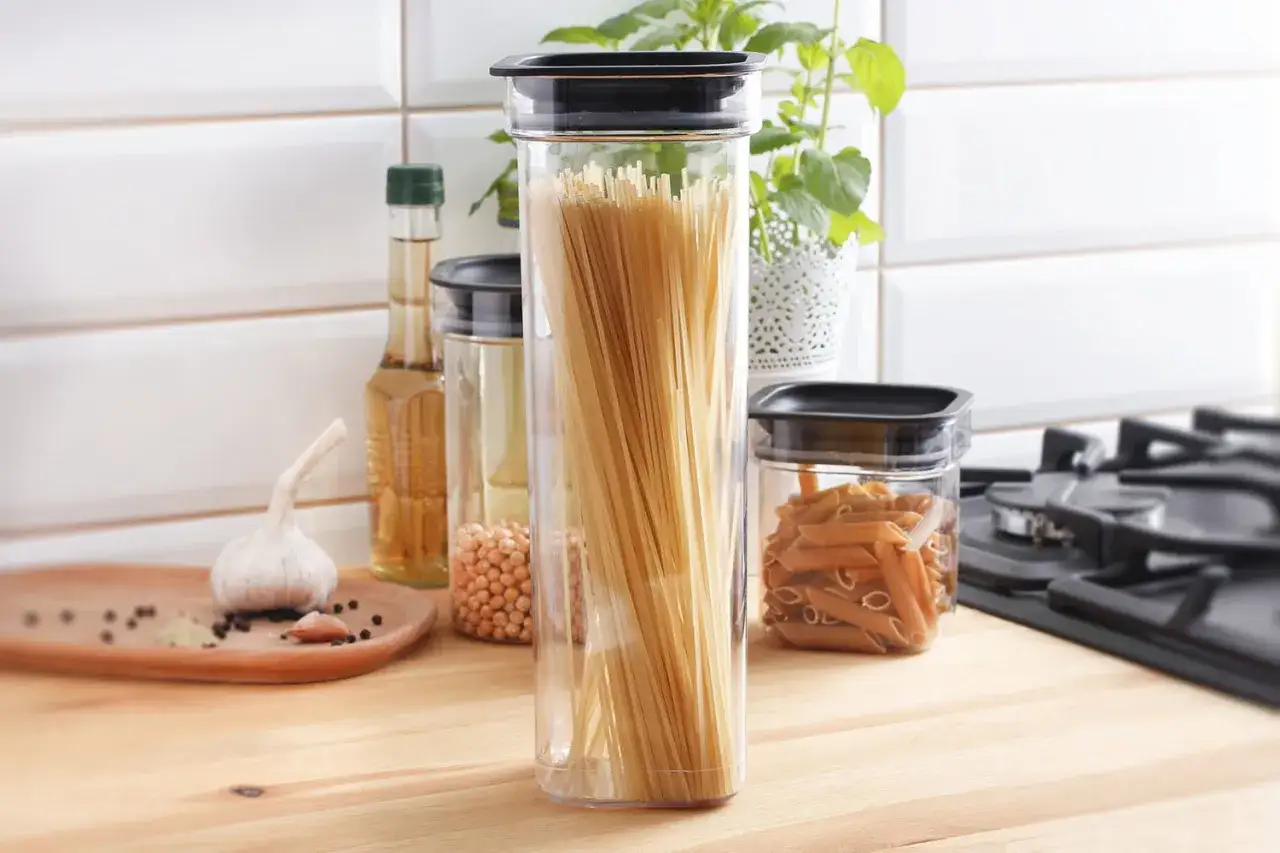 Przezroczyste pojemniki na makaron spaghetti i penne, butelki z oliwą, czosnek i przyprawy na blacie kuchennym.