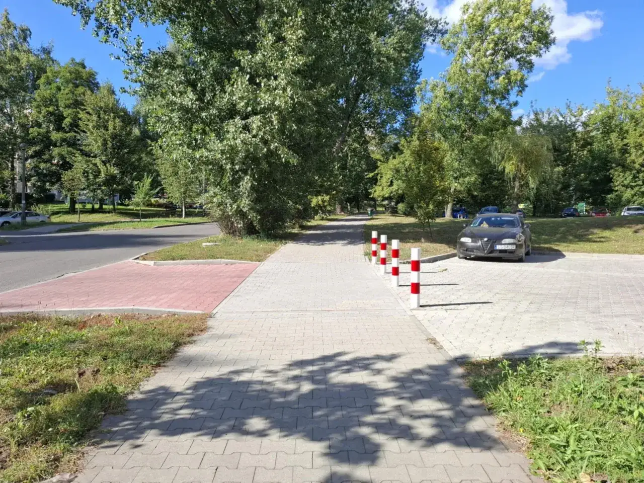Chodnik z kostki brukowej, słupki parkingowe, samochód i drzewa w słoneczny dzień.
