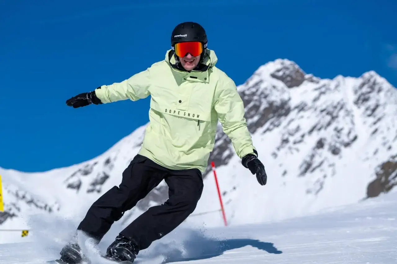 Snowboarder w jasnej kurtce i czarnych spodniach, z kaskiem i goglami, śmiga po stoku. Czy buty narciarskie pasują do snowboardu? Nie, to różne sprzęty.