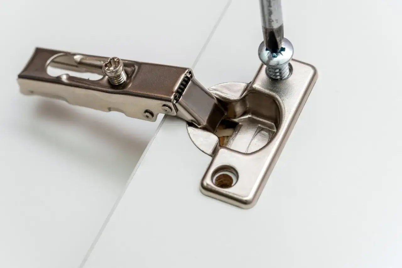 Montaż zawiasów do szafek: przykręcanie zawiasu do białej płyty meblowej za pomocą wkrętarki.