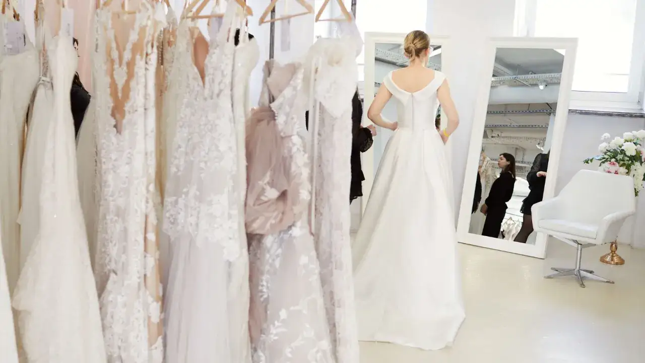 Kobieta w białej sukni ślubnej stoi przed wieszakami z sukniami i lustrem.