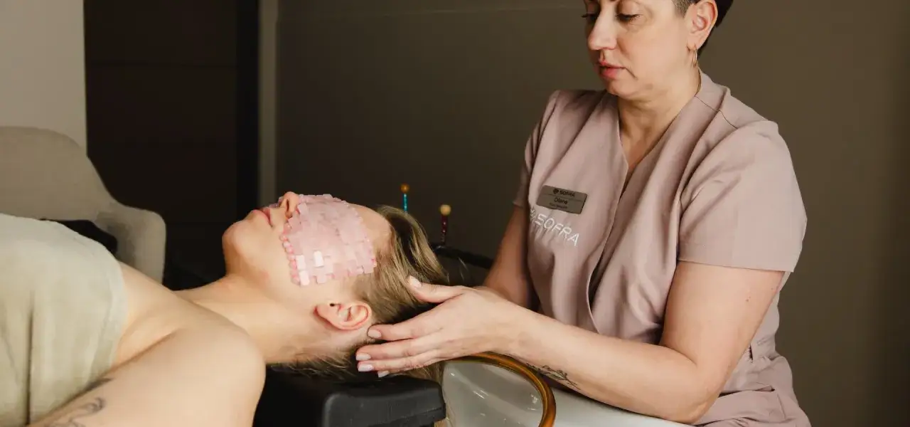 Relaks w gabinecie head spa. Kobieta leży z kamieniami na oczach, a masażystka delikatnie dotyka jej skroni.