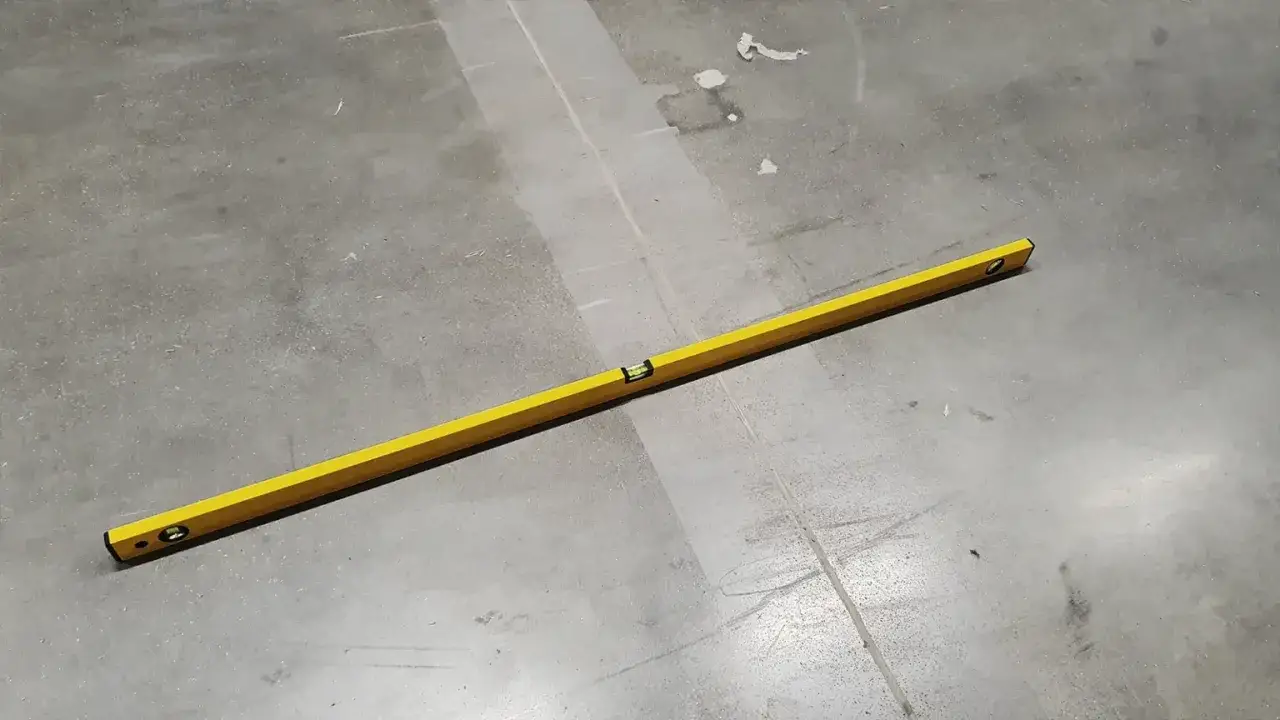 Żółta poziomica na betonowej podłodze, sprawdzająca, ile wylewki na ogrzewanie podłogowe będzie potrzebne.