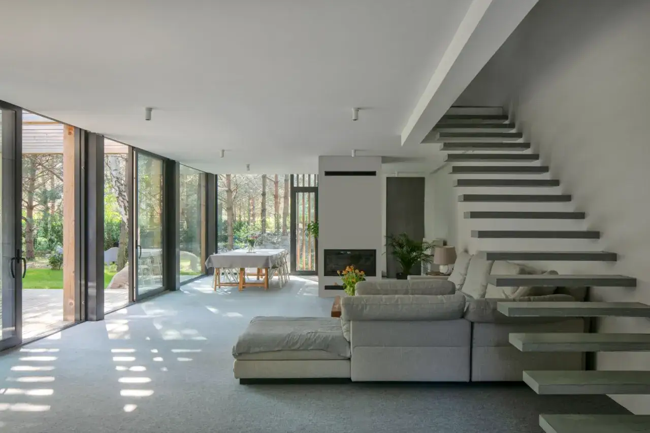 Nowoczesne świdermajer wnętrza z widokiem na las. Jasny salon z kominkiem, jadalnią i szarą sofą.