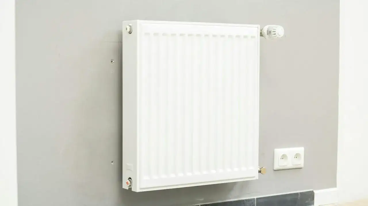 Biały grzejnik z termostatem na szarej ścianie. Dowiedz się, jak wymienić grzejnik w sezonie grzewczym, aby zapewnić ciepło w domu.