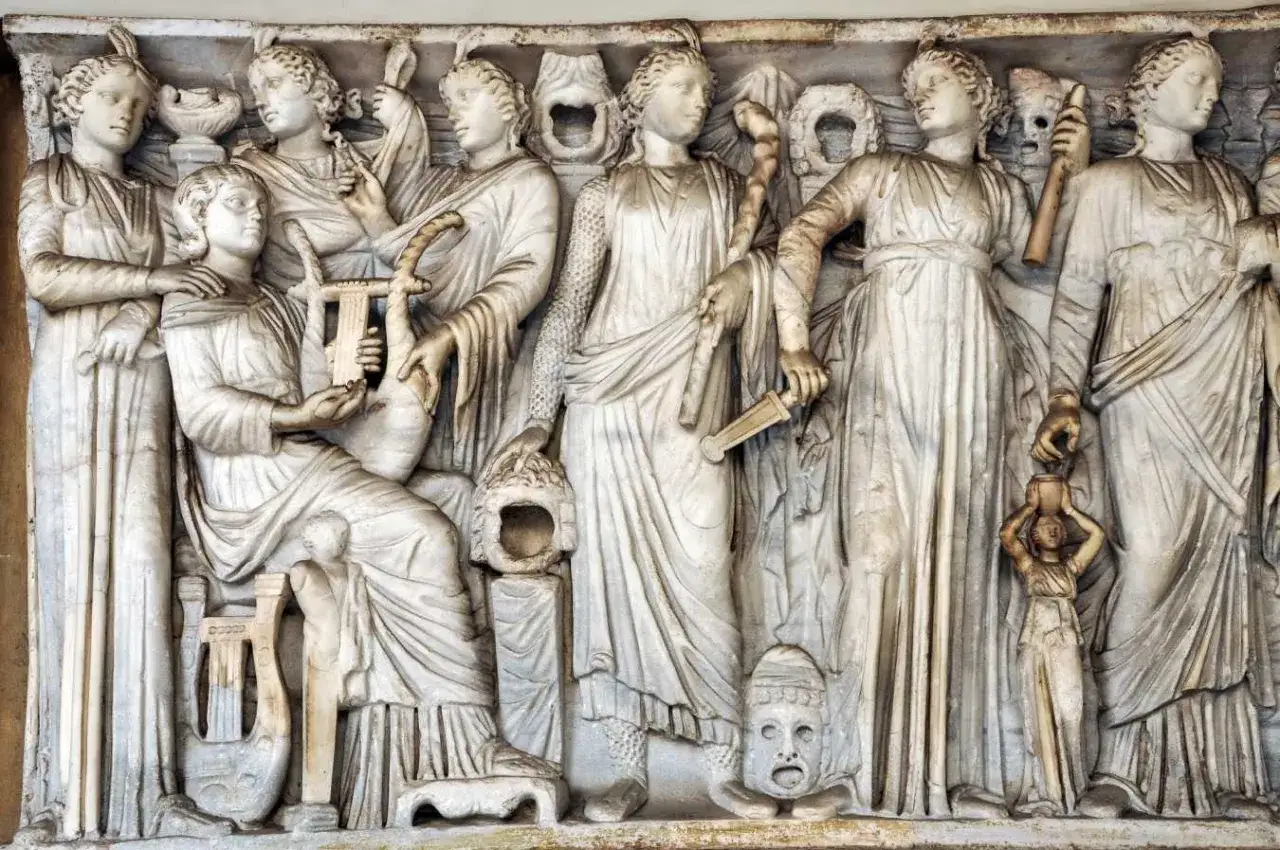 Kamienna płaskorzeźba przedstawia grupę kobiet, w tym Demeter i Korę, otoczone instrumentami muzycznymi i symbolicznymi przedmiotami.
