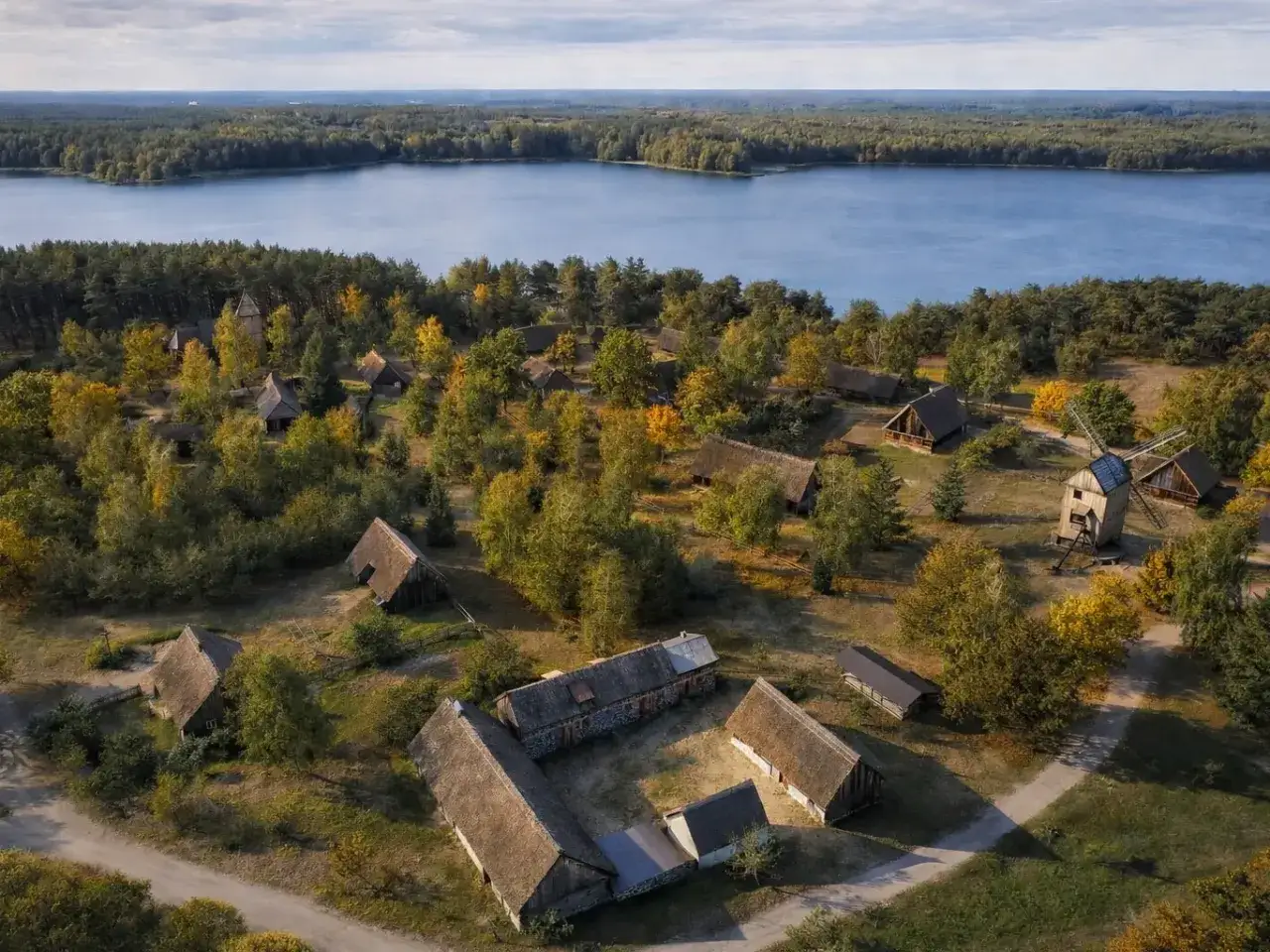 Wdzydze Kiszewskie: skansen z drewnianymi chatami i wiatrakiem nad jeziorem, otoczony jesiennym lasem.