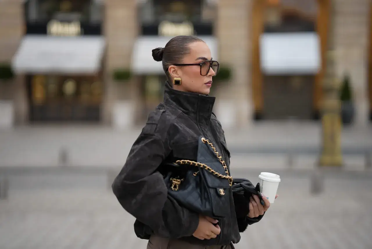 Elegancka kobieta w okularach i ciemnej kurtce puchowej, z torebką Chanel i kawą na wynos. Stylowe kurtki puchowe to podstawa jesiennych stylizacji.