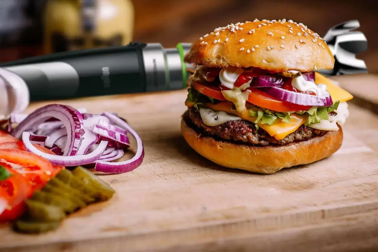 Pyszny hamburger z serem, warzywami i domowym sosem do hamburgerów. Obok pokrojona cebula i ogórki.