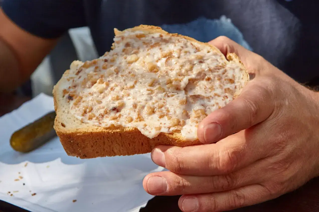 Kromka chleba posmarowana smalcem z kawałkami boczku, trzymana w dłoniach. Obok ogórek kiszony.