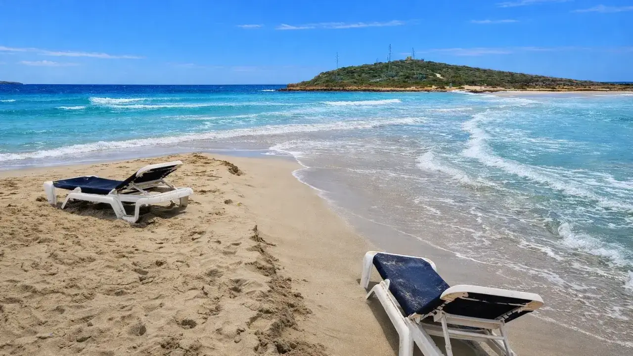 Plaża na Cyprze zimą. Dwa puste leżaki na piasku, turkusowe morze i zielone wzgórze w oddali.