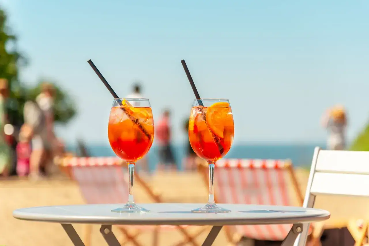 Dwa kieliszki Aperol Spritz z pomarańczą i słomką na stole, w tle plaża i morze.