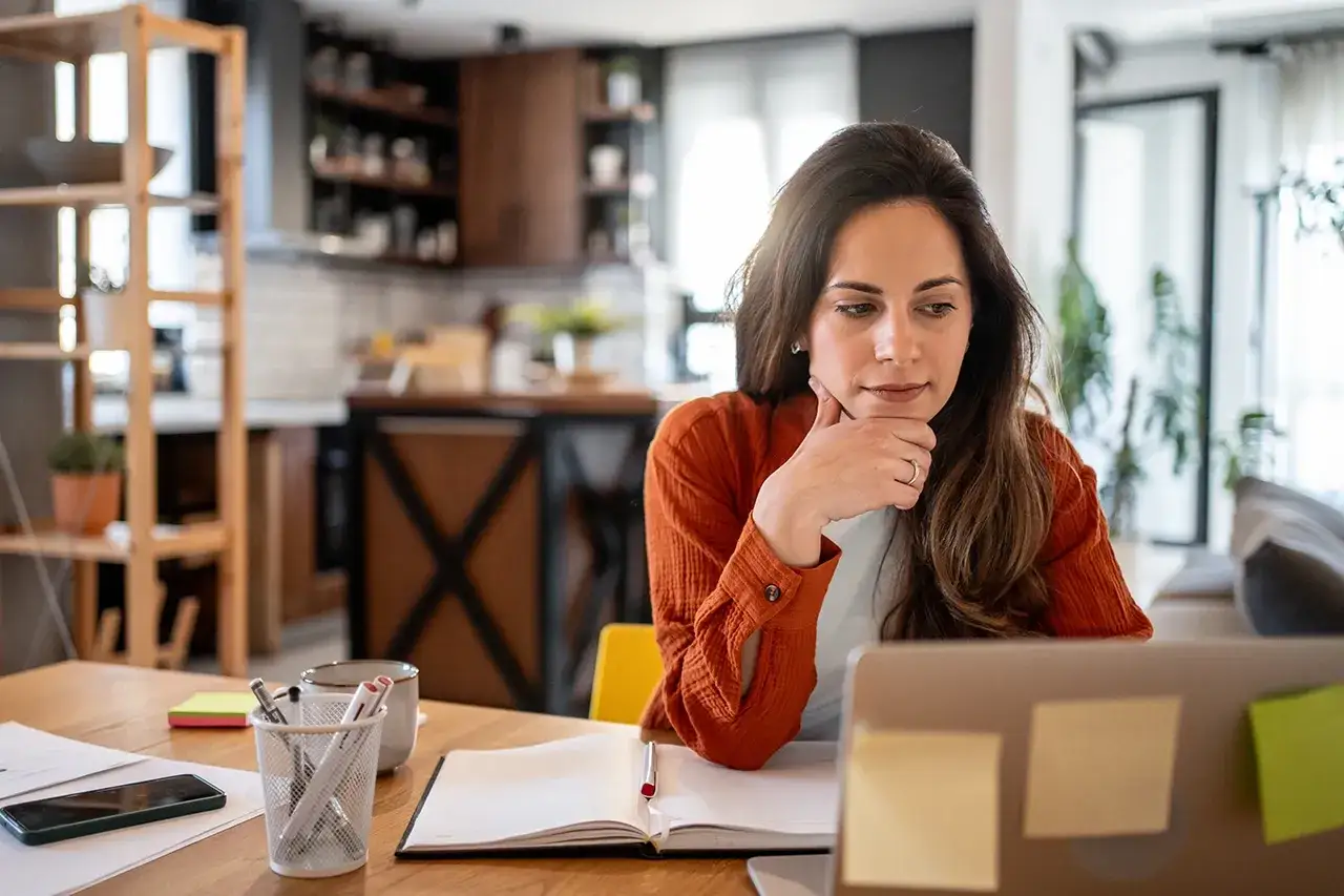 Kobieta zamyślona nad notatkami i laptopem przy biurku.